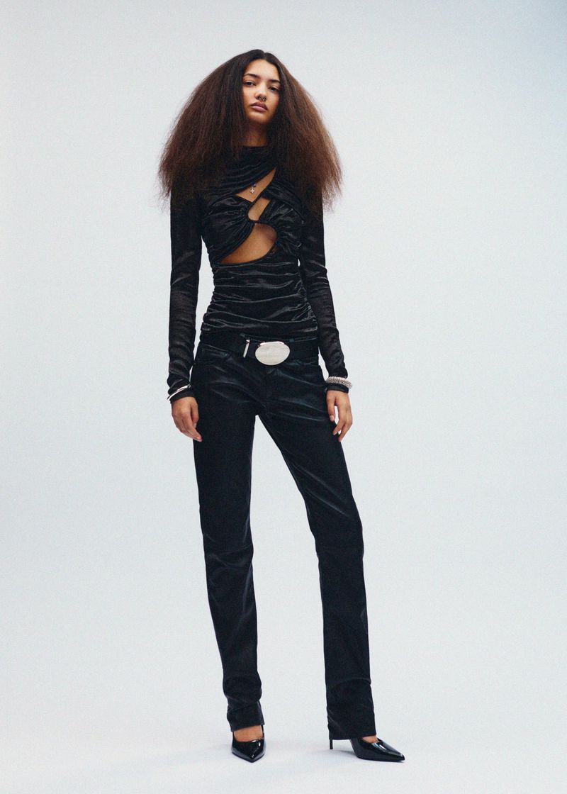 A full-length shot of a woman with voluminous brown hair, wearing a long-sleeved black top with cut-out details, and black velvet pants.