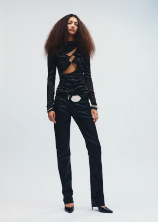 A full-length shot of a woman with voluminous brown hair, wearing a long-sleeved black top with cut-out details, and black velvet pants.