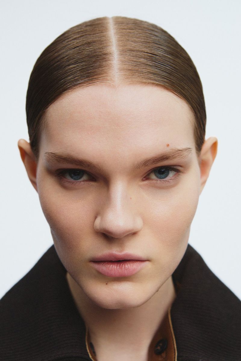 Close-up portrait of a fair-skinned young woman with slicked-back hair, blue eyes, and natural makeup wearing a brown jacket against a white background.