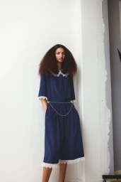 Model in a navy blue dress with lace collar standing against a plain white wall for Unpolished Magazine