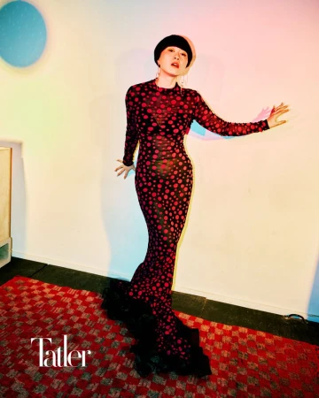 A stylish Asian woman in a red-and-black polka dot gown poses in a studio with vibrant lighting and a patterned red rug, fashion editorial.