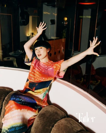 Portrait of a woman with bowl cut, wearing a colorful pleated outfit, sitting on a white sofa with brown cushions, with hands raised.