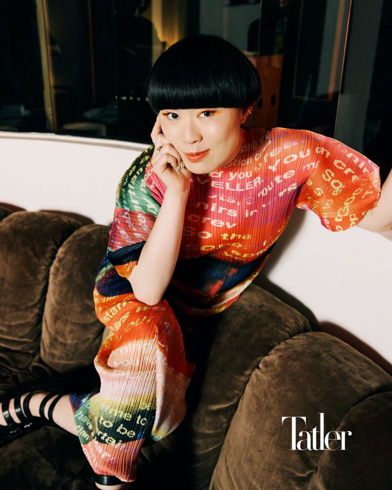 Portrait of an Asian woman with a bowl cut wearing a multicoloured, text-printed shirt, sitting on a brown sofa in an indoor setting.