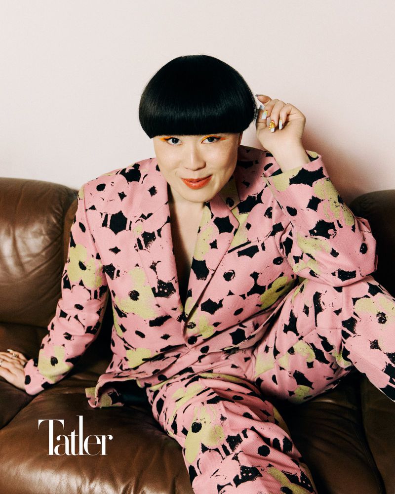 A woman with a dark bowl cut, wearing a pink floral suit, is seated on a brown leather sofa indoors in a fashion forward portrait.