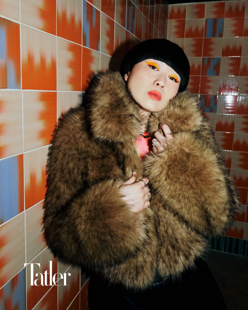 Adult woman with vibrant makeup poses indoors wearing a fur coat with a black hat against an orange and blue tile wall.