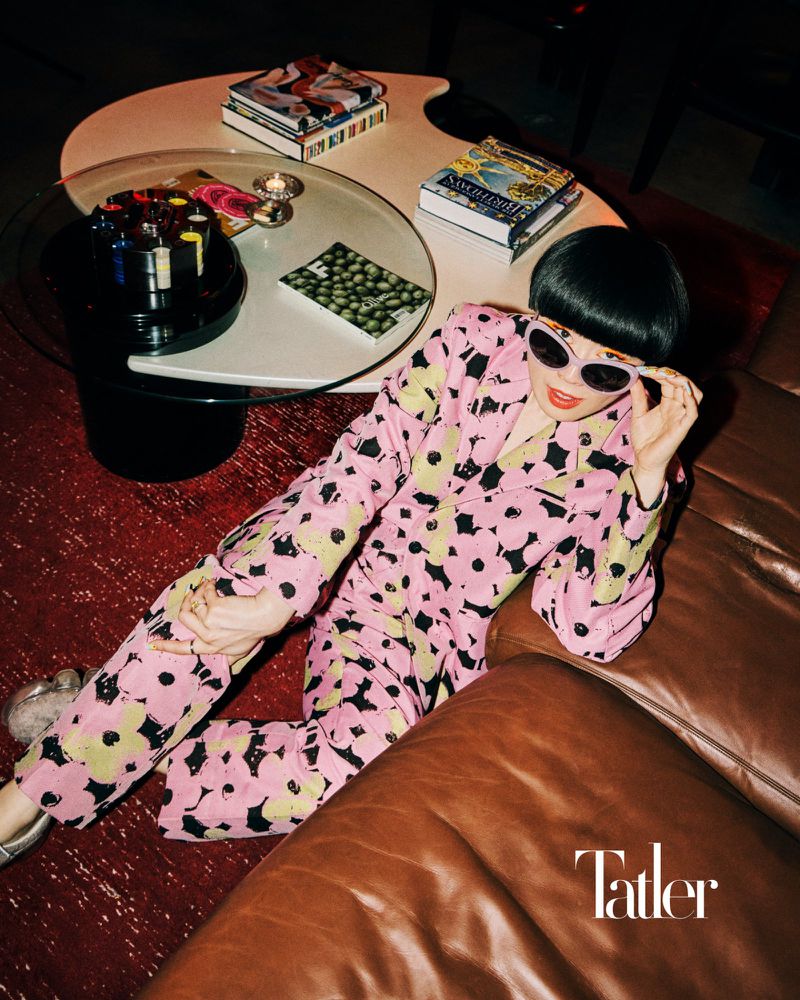 Woman in floral pyjamas poses indoors next to a glass table and a brown leather couch, illuminated by ambient lighting, fashion editorial.
