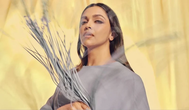 Close-up portrait of an adult woman with shoulder-length hair, holding tall gray grass, set against a yellow background with soft lighting.