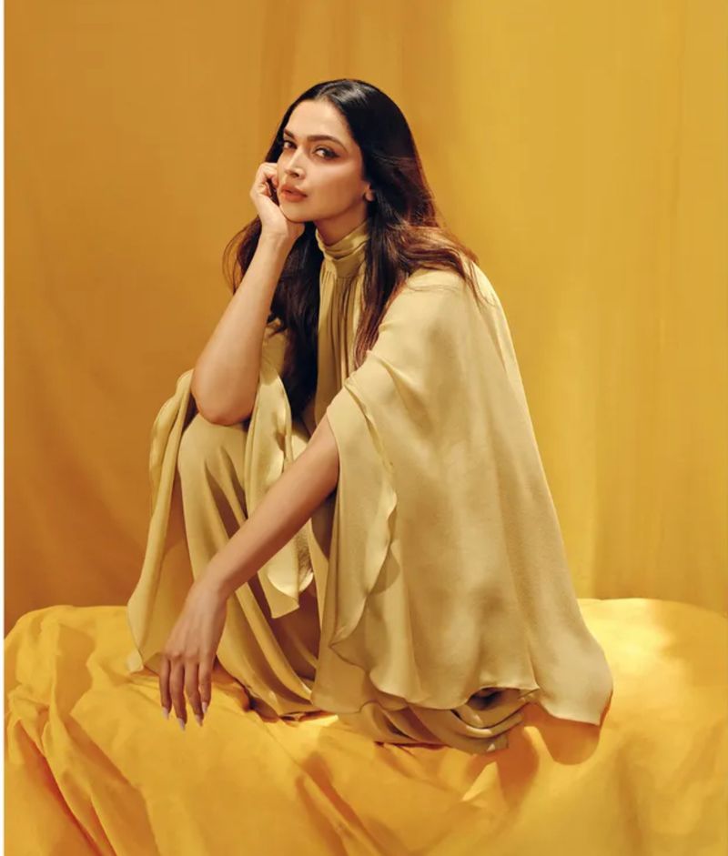 A woman with long dark hair poses on a golden yellow backdrop wearing a high-necked silky yellow dress with flowing cape sleeves.