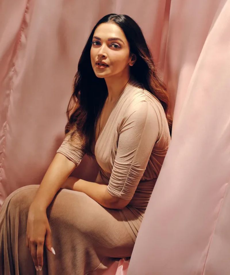 Portrait of a South Asian woman with dark hair wearing a beige dress posing in front of a pink fabric backdrop in a photo studio.