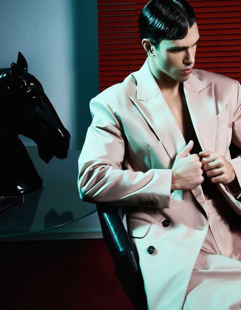 A stylish adult male model in a beige suit poses in a dimly lit room next to a black horse head statue for a fashion editorial.