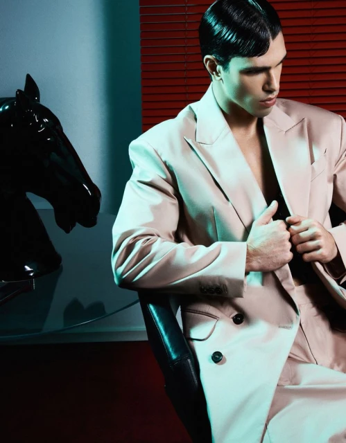A stylish adult male model in a beige suit poses in a dimly lit room next to a black horse head statue for a fashion editorial.