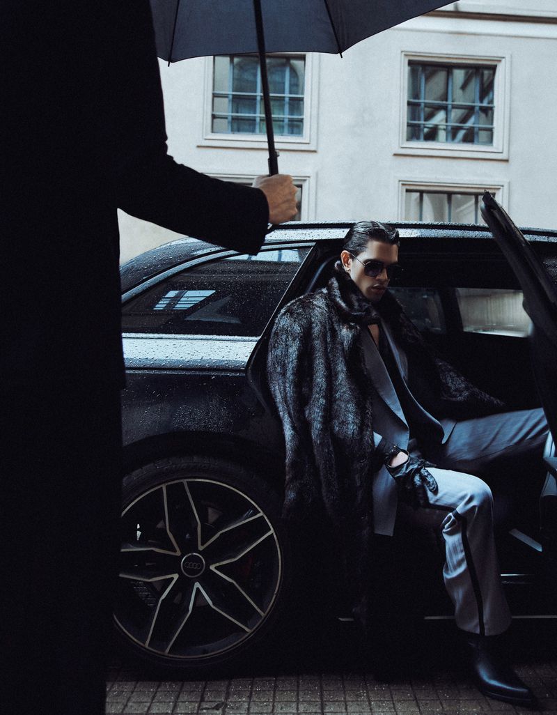 Stylish man in suit and fur coat exits black car under umbrella on city street. Fashion shoot with urban backdrop.