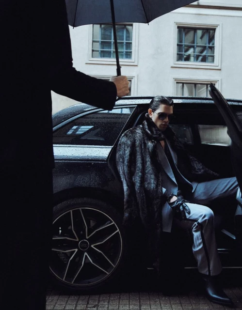 Stylish man in suit and fur coat exits black car under umbrella on city street. Fashion shoot with urban backdrop.