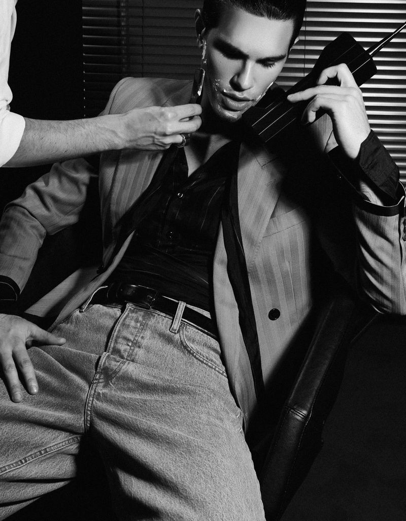 Black and white portrait of a man having his face shaved by another man while holding a large vintage cell phone in an indoor setting.