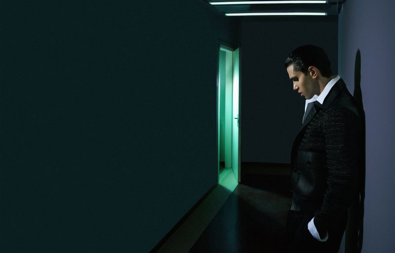 Stylish man in suit leans against wall in a dimly lit corridor with modern lighting, a door ajar, and a moody, contemplative atmosphere.