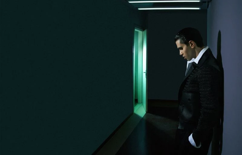 Stylish man in suit leans against wall in a dimly lit corridor with modern lighting, a door ajar, and a moody, contemplative atmosphere.