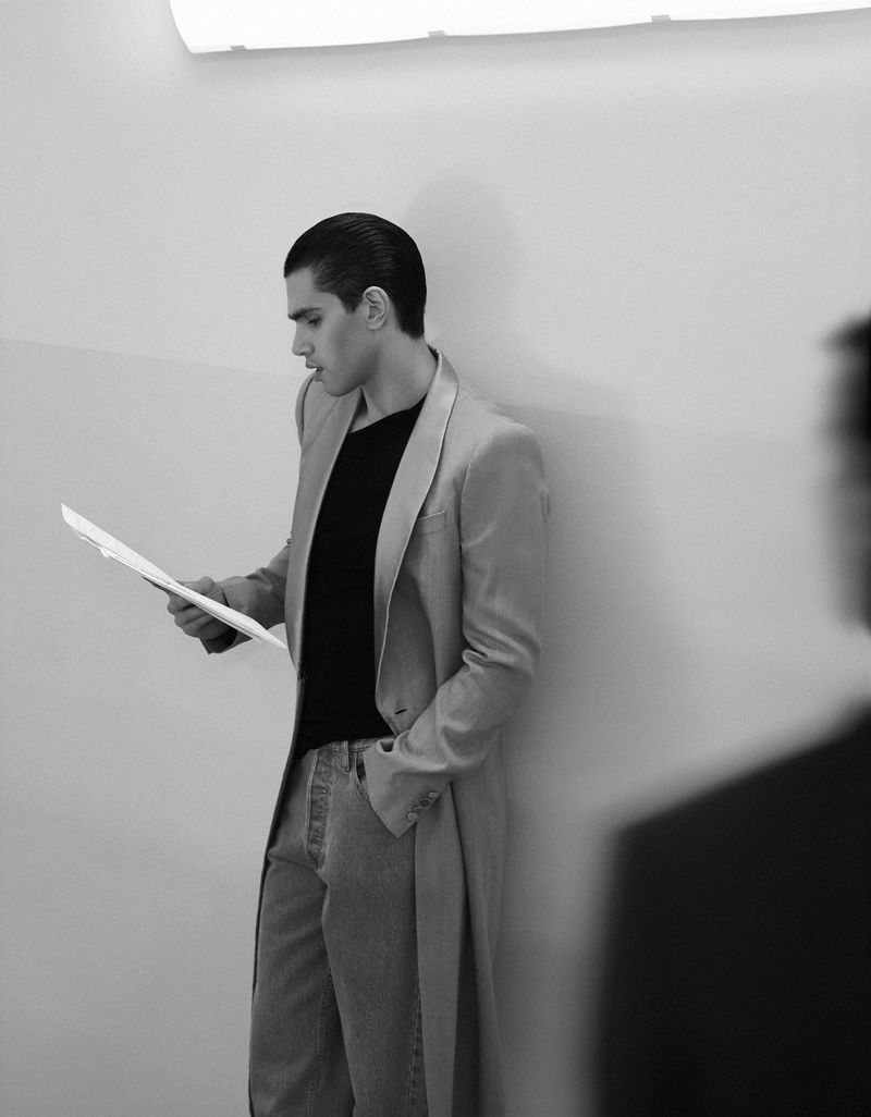 Monochrome portrait of a young adult man in a suit and jeans, holding papers and standing against a wall with soft studio lighting.