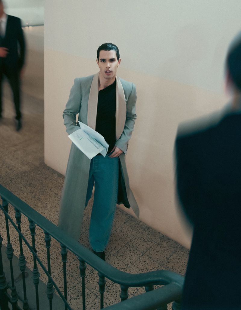 An adult man stands on a staircase in an indoor setting, holding papers in his hand and wearing a long coat and casual pants.