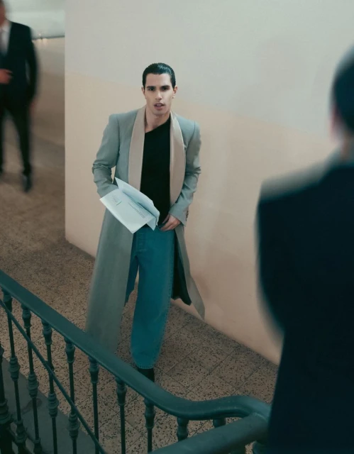 An adult man stands on a staircase in an indoor setting, holding papers in his hand and wearing a long coat and casual pants.