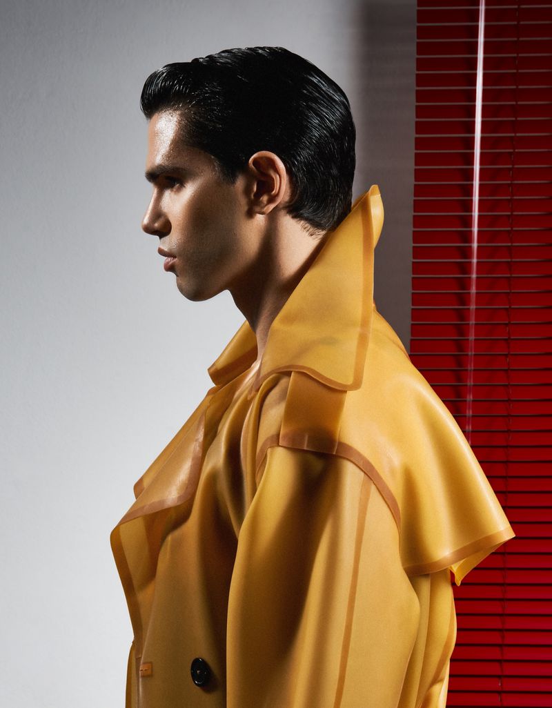 Stylish man in yellow vinyl trench coat poses in studio against a red louvered wall. Fashion, design, and portrait photography.
