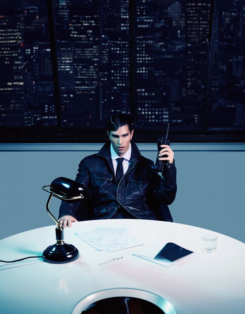 Stylish man in a black jacket, sitting at a futuristic white desk with a city skyline view, holding a walkie-talkie. Editorial fashion.