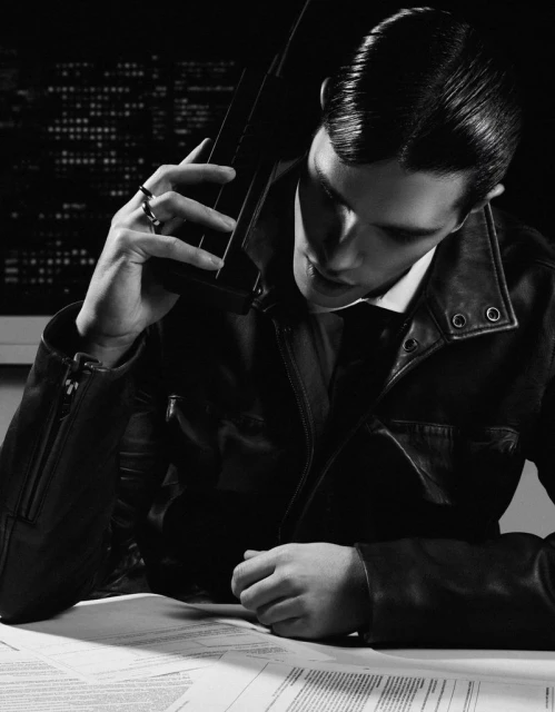 Monochrome close-up of a man in a leather jacket talking on a vintage telephone, seated at a desk with papers, set against a city lights backdrop.
