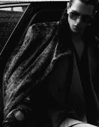 Monochrome portrait of a male model wearing dark sunglasses and a furry coat exiting a car with rain droplets.