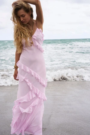 Woman in flowing pink dress poses on beach with ocean waves in the background. Long blonde hair, jewelry, serene expression, overcast sky.