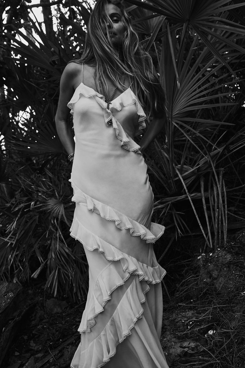 Woman in ruffled gown stands amongst palm fronds in black-and-white, showcasing fashion in a natural setting with tropical foliage.