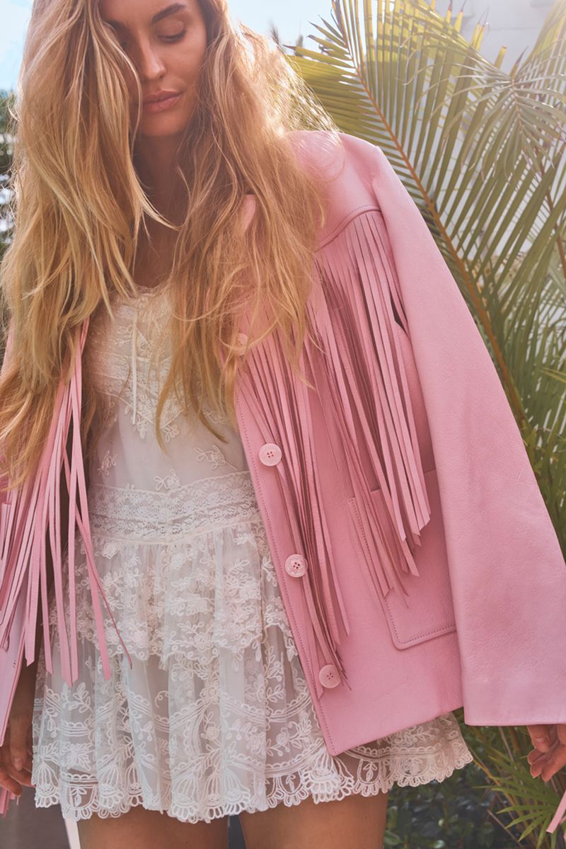 Woman in fringed pink jacket and white lace dress poses outdoors with palm trees. Fashion, sunshine, natural light.