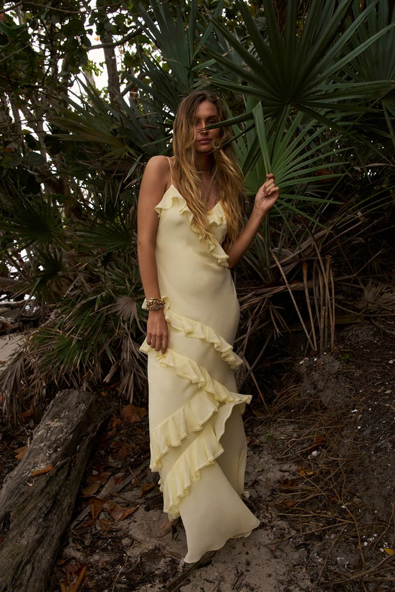 A woman in a yellow dress poses among palm trees in a natural setting. She has long hair and is wearing a long, flowing dress.