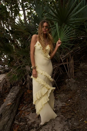A woman in a yellow dress poses among palm trees in a natural setting. She has long hair and is wearing a long, flowing dress.