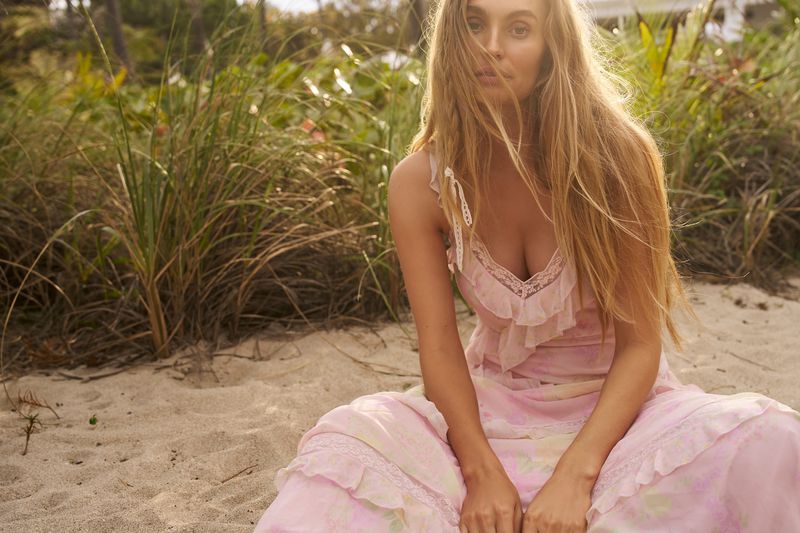 A blonde woman in a pink dress sits on a sandy beach with native grasses in the background in a fashion or beauty editorial shoot.