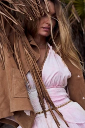 Model wearing a fringed brown jacket over a pink and white dress amid foliage