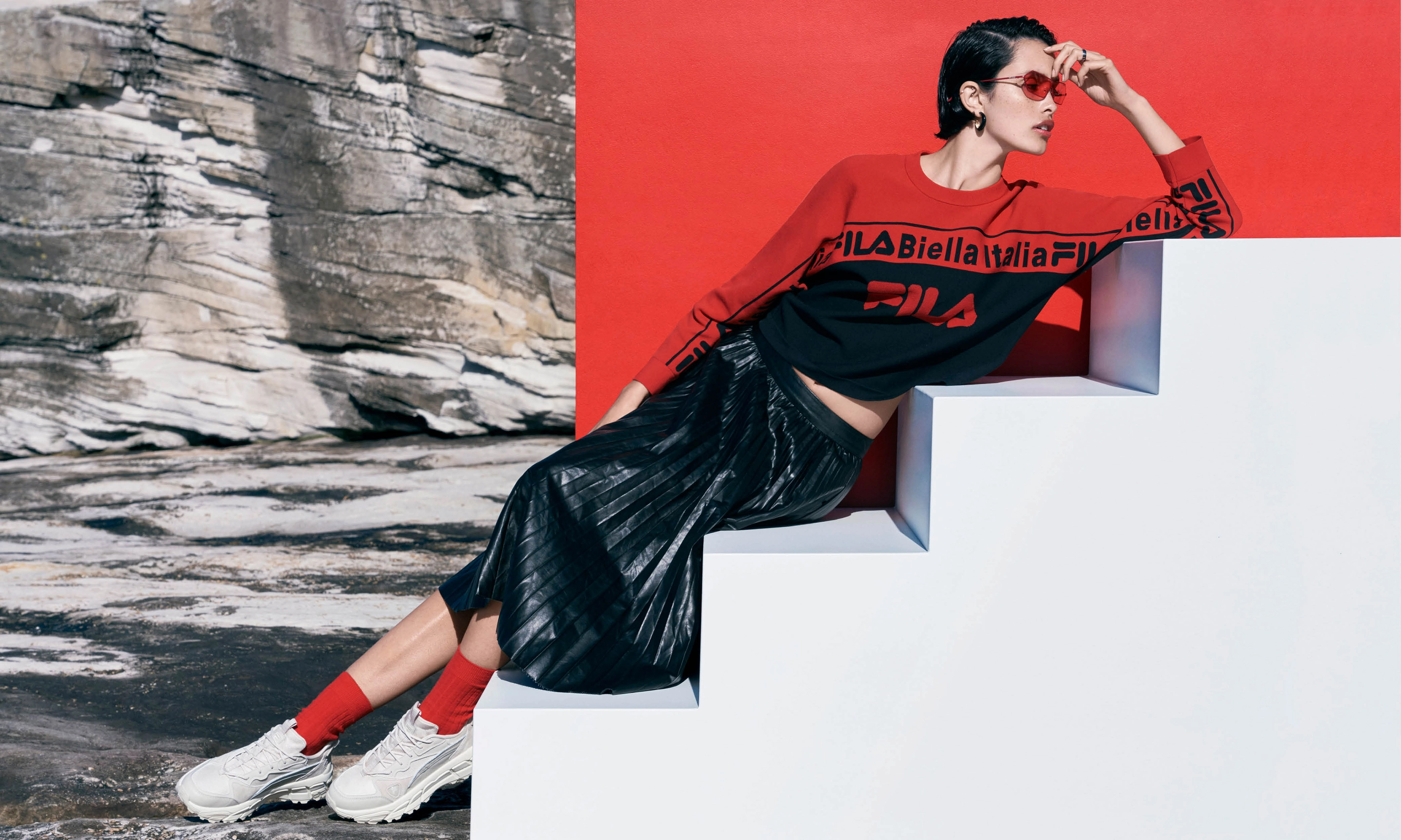 Woman in FILA sweater and skirt poses on white steps against a split red wall and rocky outdoor scene, fashion editorial style.