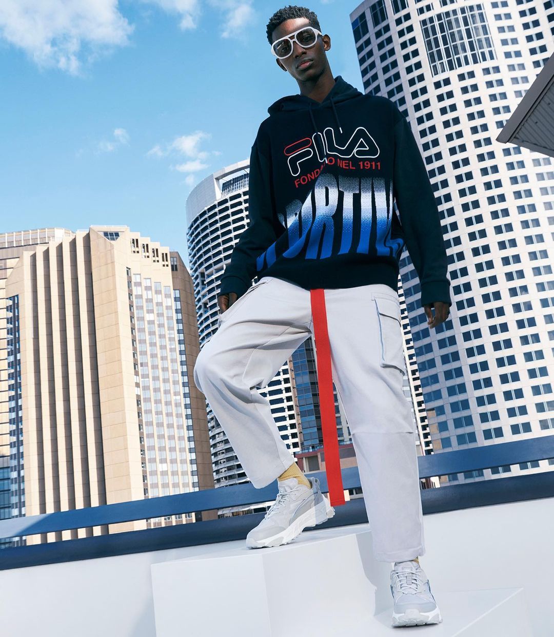 Fashion shoot of a Black male model wearing a Fila hoodie, pants, and sneakers, posing against a city skyline backdrop on a rooftop.