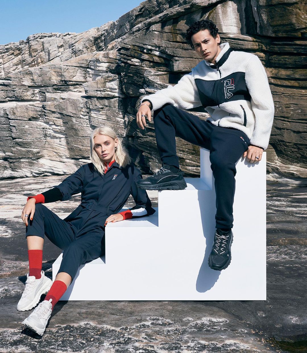 Two fashion models in athletic wear pose on white block stairs with a rocky mountain backdrop under a bright blue sky.