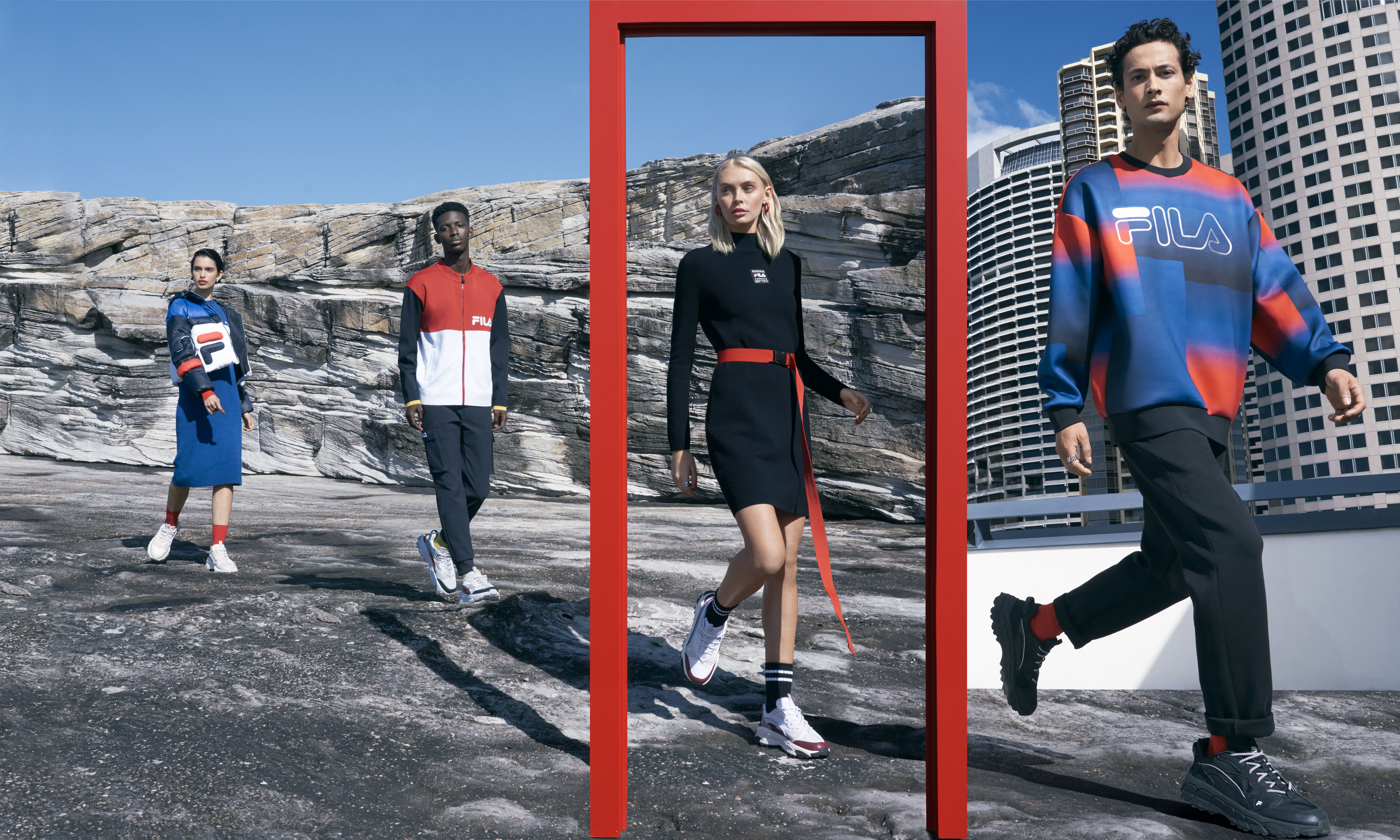 Four models wearing Fila apparel stand on a concrete surface with a rocky hillside and city skyline in the background on a sunny day.
