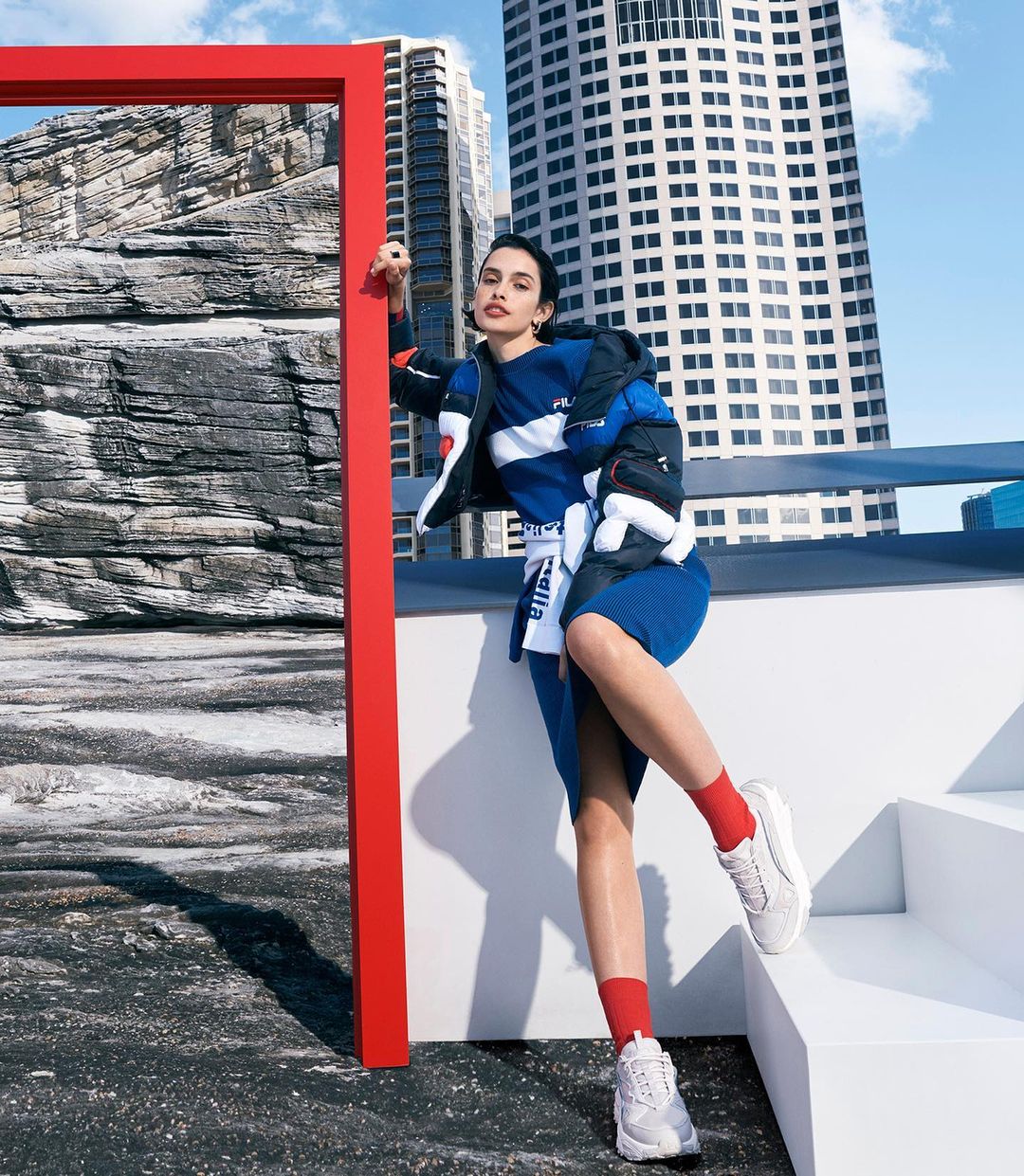 A stylish woman poses outdoors in a blue dress and white sneakers, with a modern red frame and city buildings in the backdrop.