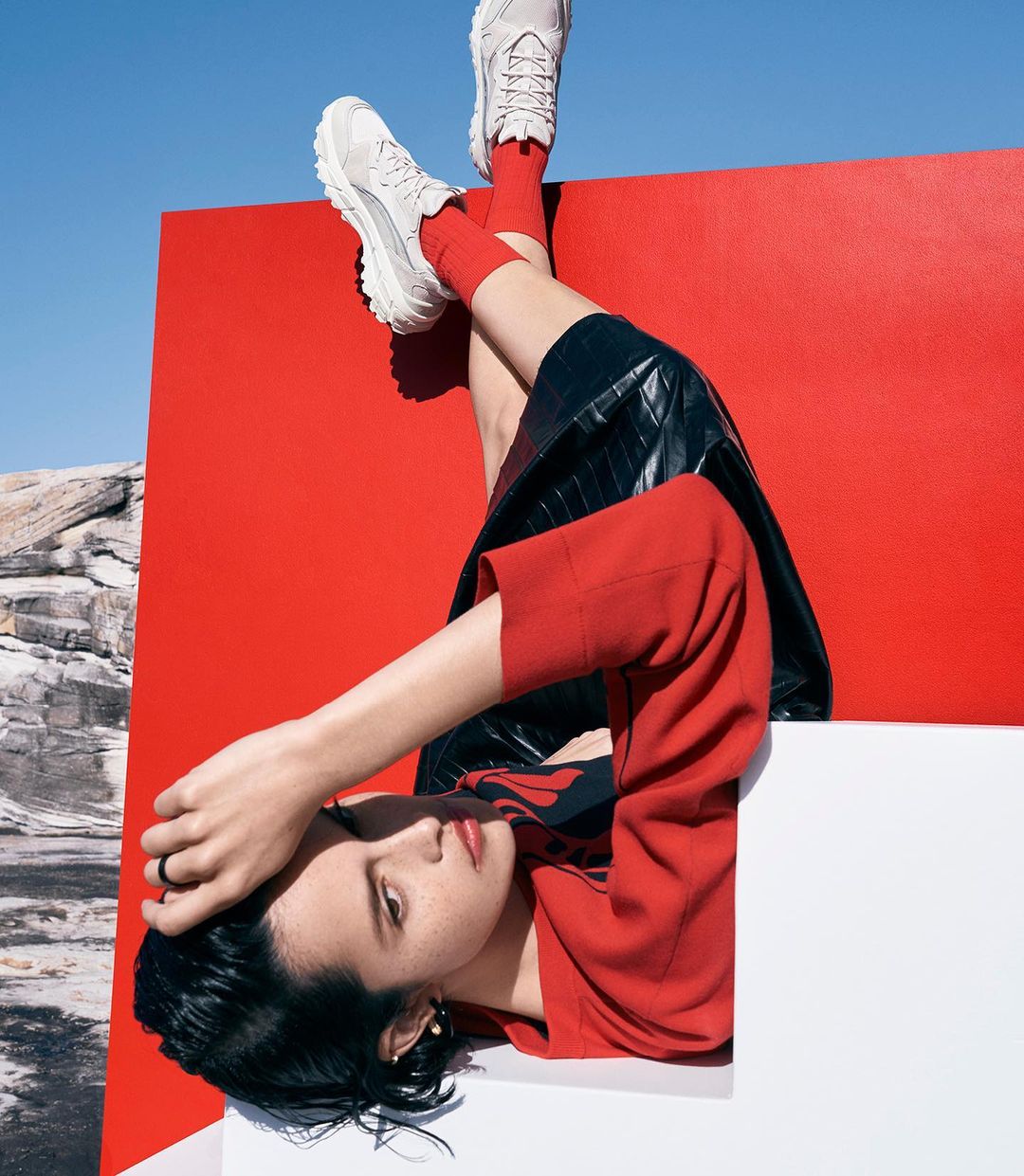 Upside down fashion shot of a brunette woman in red top and sneakers, against a red backdrop and blue sky, with rock formations.
