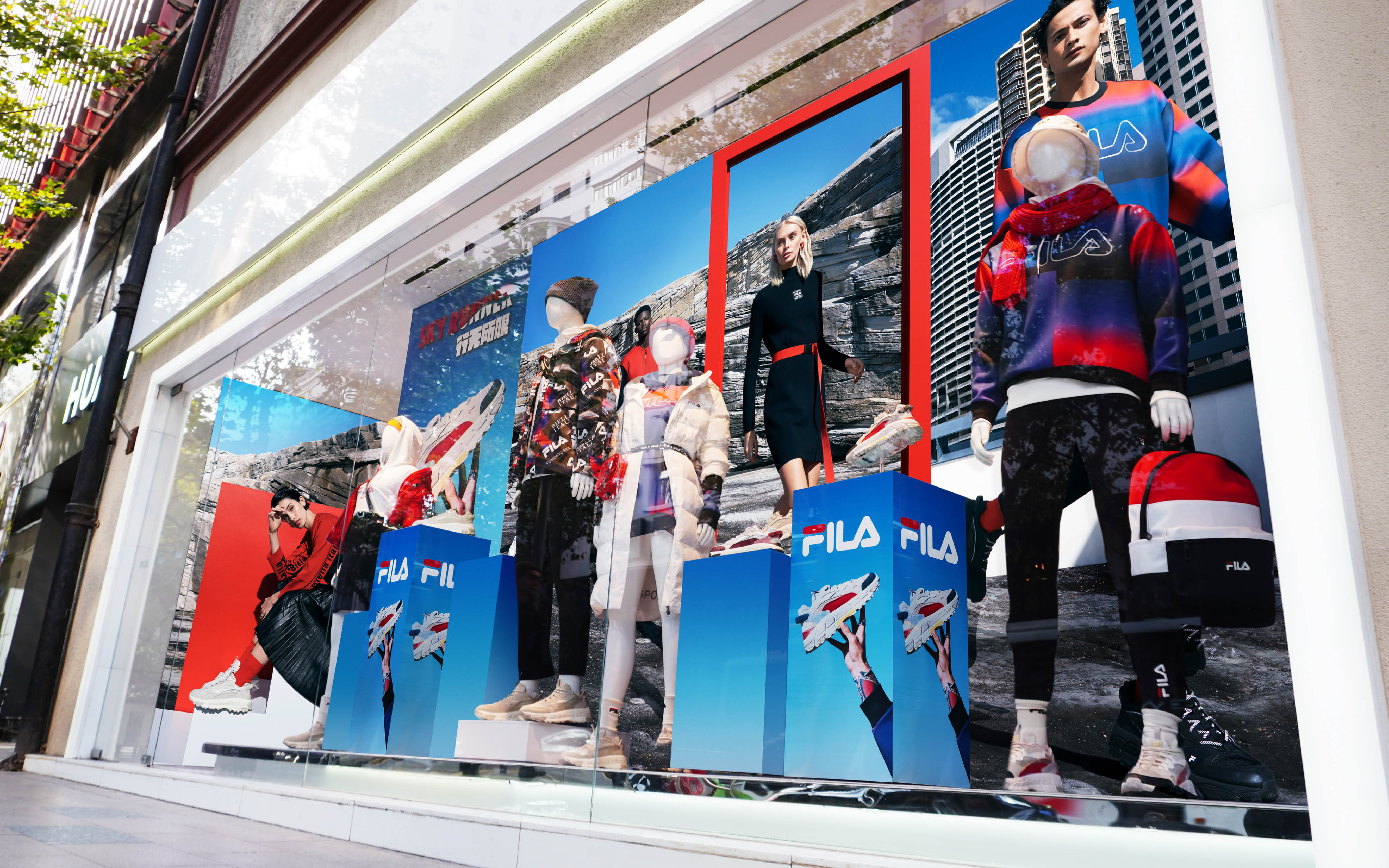 Storefront window display features mannequins in FILA sportswear against a backdrop of mountain and city images, showcased on blue blocks.