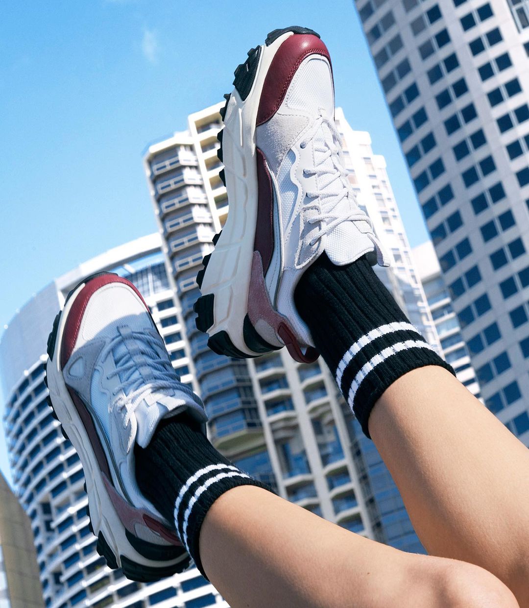 Close-up of white and maroon athletic shoes worn with striped socks against a backdrop of tall city buildings and bright blue sky.