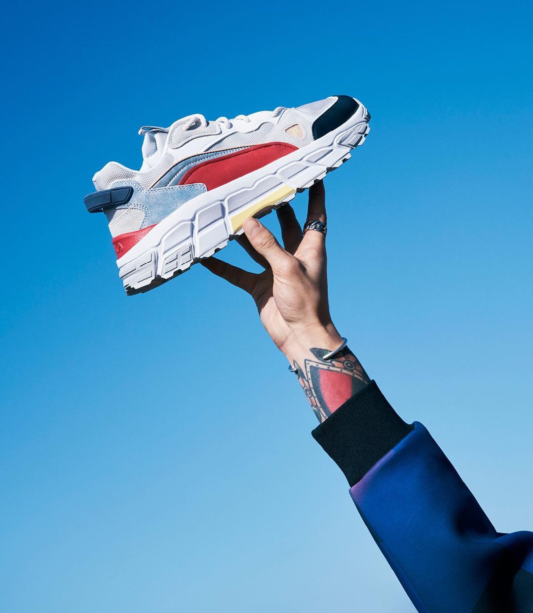 A single running shoe is held up in a hand against a blue sky background. The athletic shoe is red, white, and black.