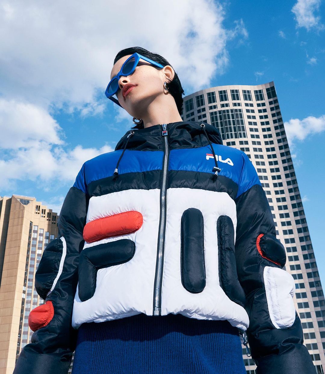 Woman in a Fila puffer jacket, blue sunglasses, and city backdrop. Fashionable outerwear in an urban setting against a bright blue sky.