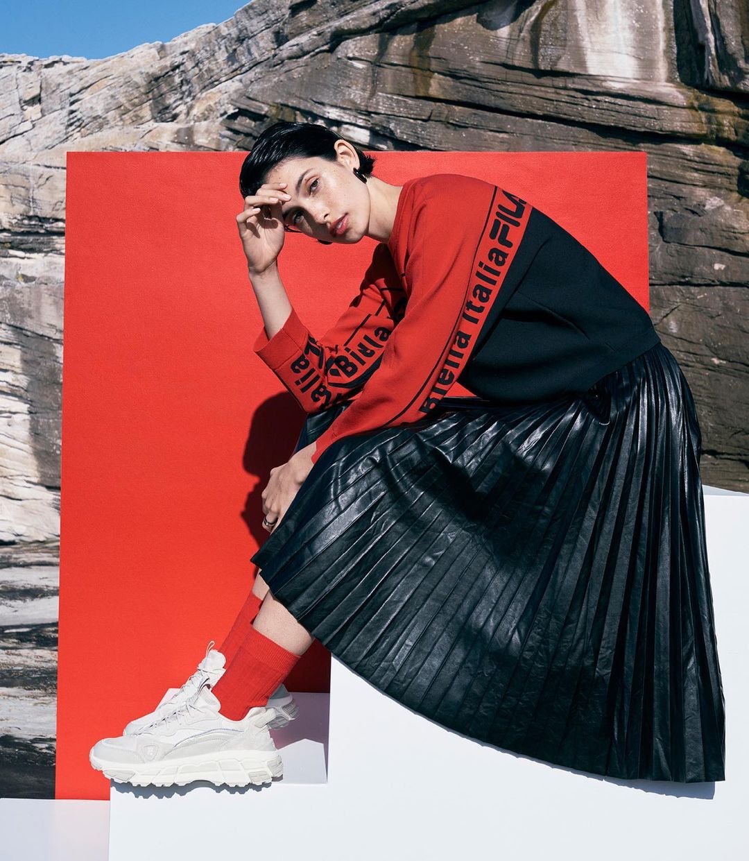A woman in athletic clothing sitting with neutral expression against a red panel, rocks, and blue sky; wearing athletic shoes.