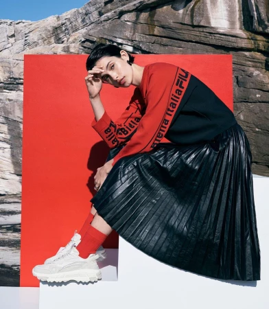 A woman in athletic clothing sitting with neutral expression against a red panel, rocks, and blue sky; wearing athletic shoes.