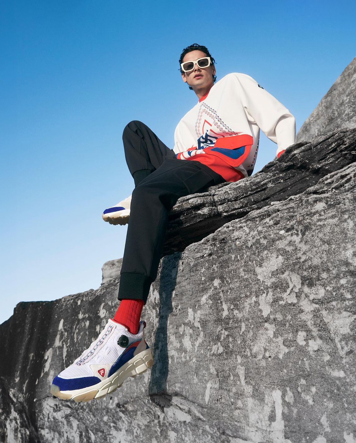 Man sitting on a rock wearing a sweater, pants, sunglasses, and sneakers, posing with a clear blue sky in the background.