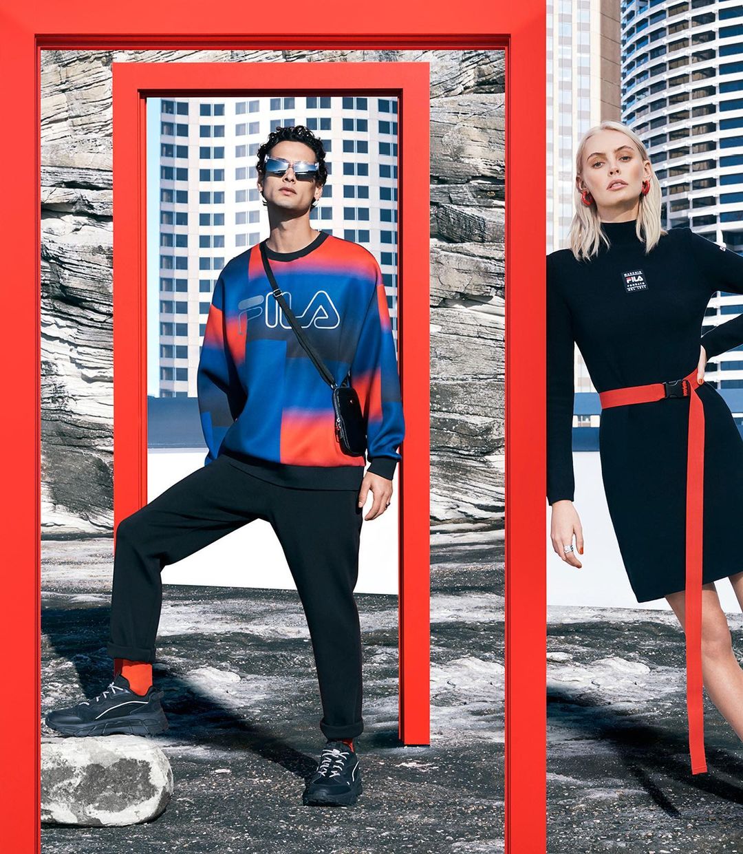 Two fashion models pose outdoors in Fila apparel; a man wears a color-blocked sweatshirt, and a woman wears a little black dress with a red belt.
