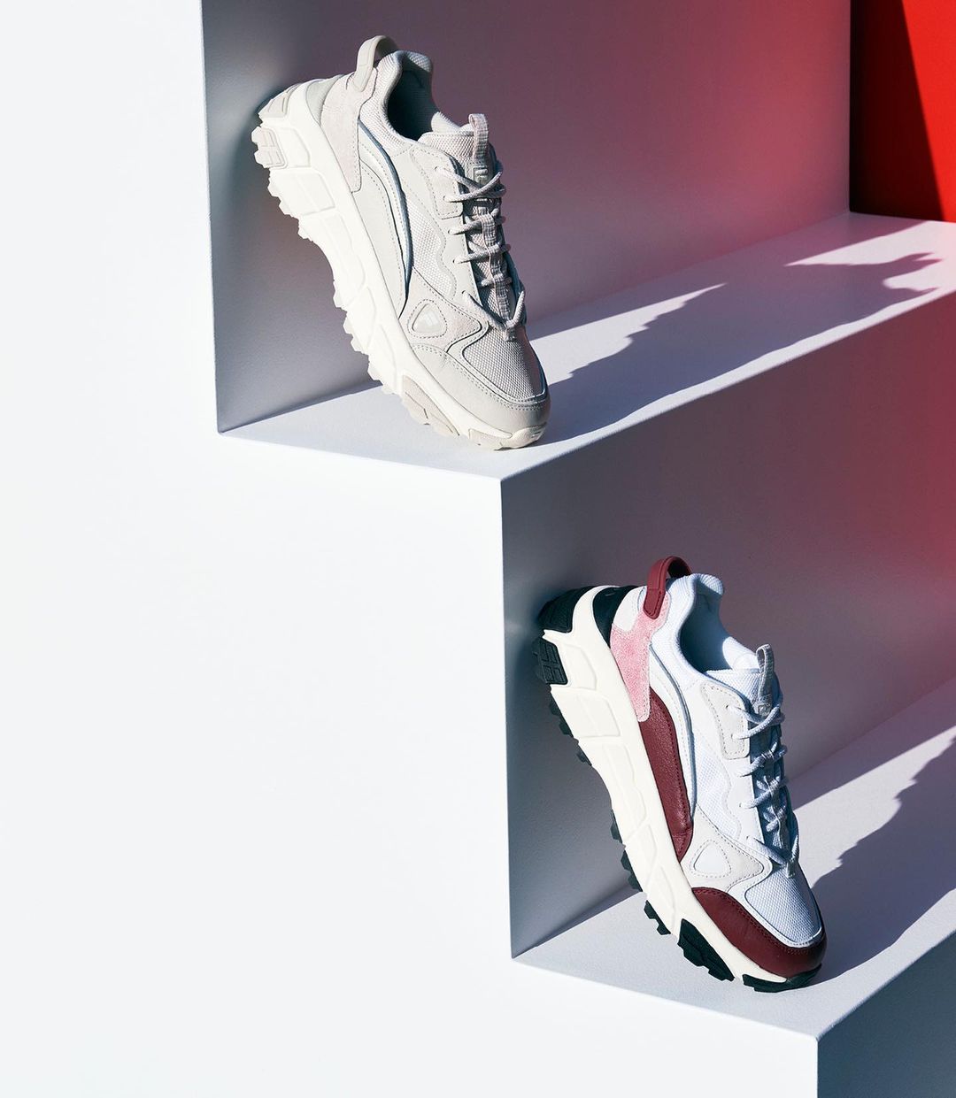 Two athletic shoes, one grey and one white and burgundy, displayed on angled white steps against a white and red gradient background.