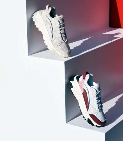 Two athletic shoes, one grey and one white and burgundy, displayed on angled white steps against a white and red gradient background.